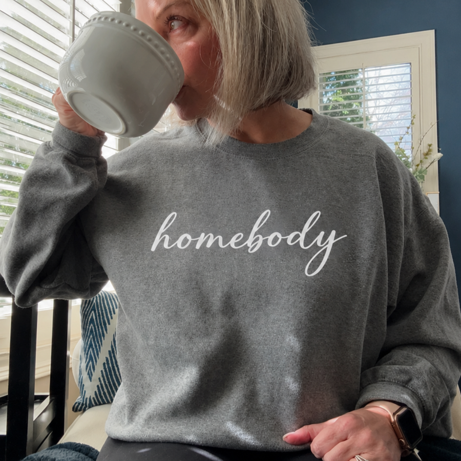 A woman in a gray sweatshirt that says "homebody" drinking coffee