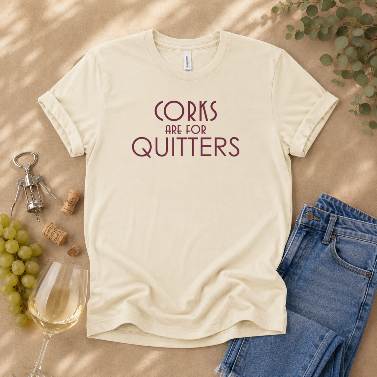 A cream-colored t-shirt with the text 'CORKS ARE FOR QUITTERS' printed in maroon, styled in a flat lay with wine glasses, grapes, a corkscrew, and blue jeans on a tan background.