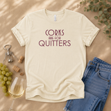 A cream-colored t-shirt with the text 'CORKS ARE FOR QUITTERS' printed in maroon, styled in a flat lay with wine glasses, grapes, a corkscrew, and blue jeans on a tan background.