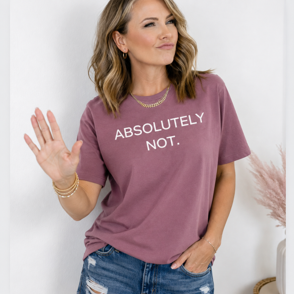 A woman wearing a dusty rose short-sleeve t-shirt with the text 'ABSOLUTELY NOT.' printed in white across the chest, paired with distressed blue jeans and gold jewelry, posing against a light gray wall.