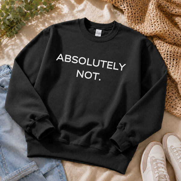 A black crewneck sweatshirt with the text 'ABSOLUTELY NOT.' printed in white across the chest, styled in a flat lay with jeans, a woven blanket, white sneakers, and green leaves on a beige fabric background.