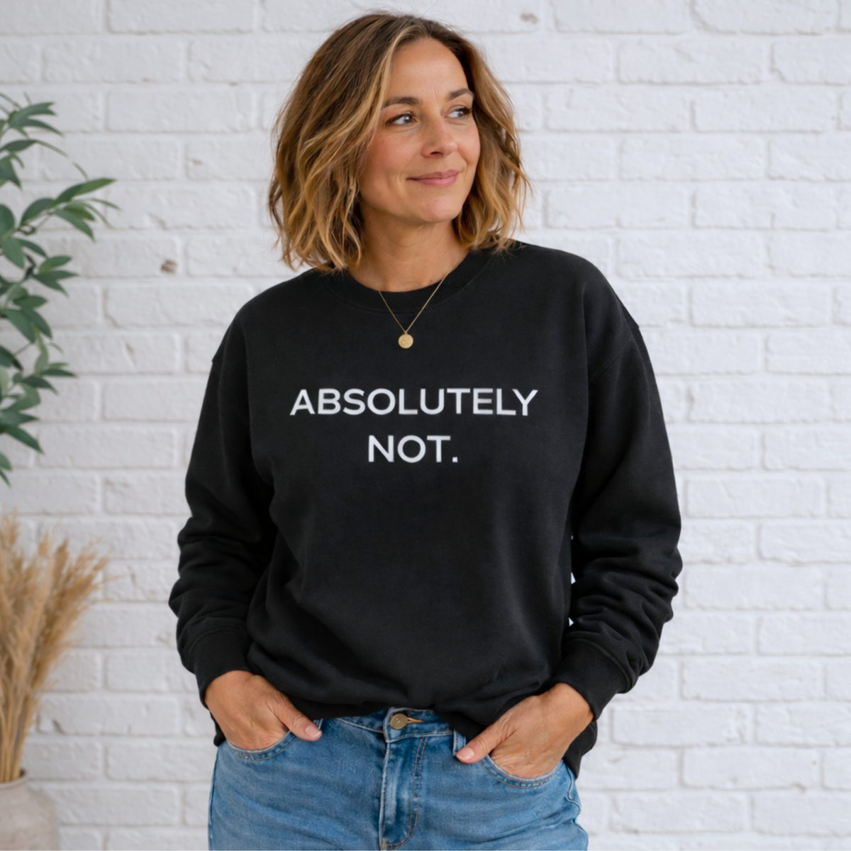 A woman standing in a black sweatshirt with the words "Absolutely Not" in white text and jeans, casually leaning in front of a brick wall.