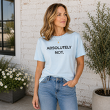 A woman wearing a light blue graphic t-shirt with the text 'ABSOLUTELY NOT.' printed in black, paired with blue jeans, standing in front of a white brick wall with potted plants.