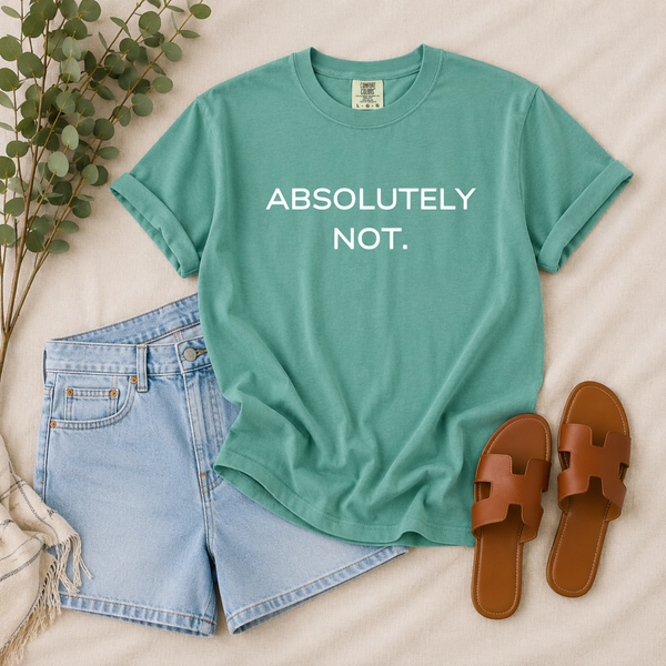 A styled flat-lay featuring a sage green t-shirt with the text 'ABSOLUTELY NOT.' in white, paired with light blue denim shorts and brown slide sandals on a beige fabric background with eucalyptus branches.