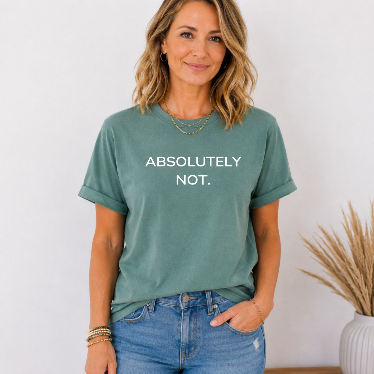 A woman wearing a sage green graphic t-shirt with the text 'ABSOLUTELY NOT.' printed in white across the chest, paired with blue jeans, standing against a light wall with a potted plant in the background.