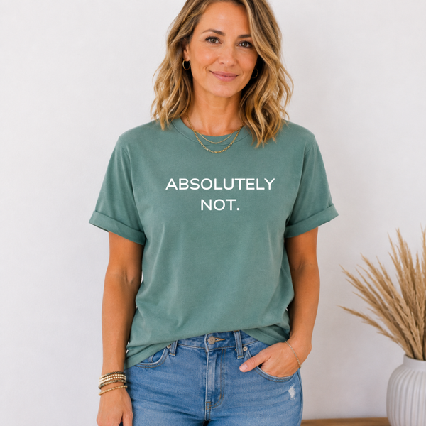 A woman wearing a sage green graphic t-shirt with the text 'ABSOLUTELY NOT.' printed in white across the chest, paired with blue jeans, standing against a light wall with a potted plant in the background.