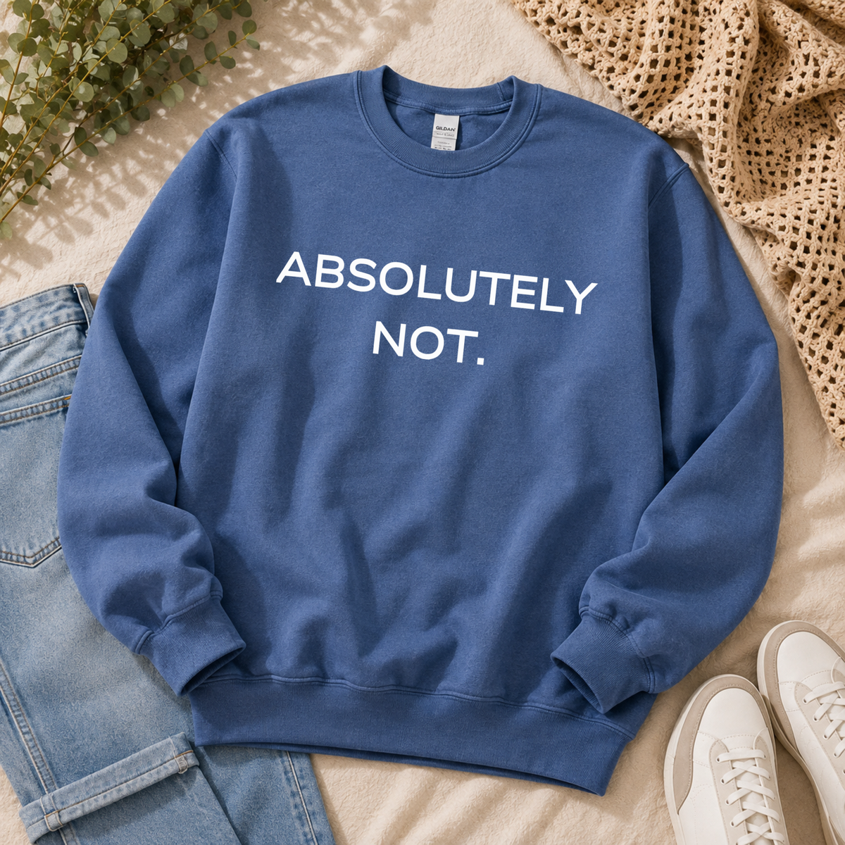 A blue crewneck sweatshirt with the text 'ABSOLUTELY NOT.' printed in white across the chest, styled in a flat lay with light-wash jeans, white sneakers, a beige woven blanket, and green foliage on a light beige background.