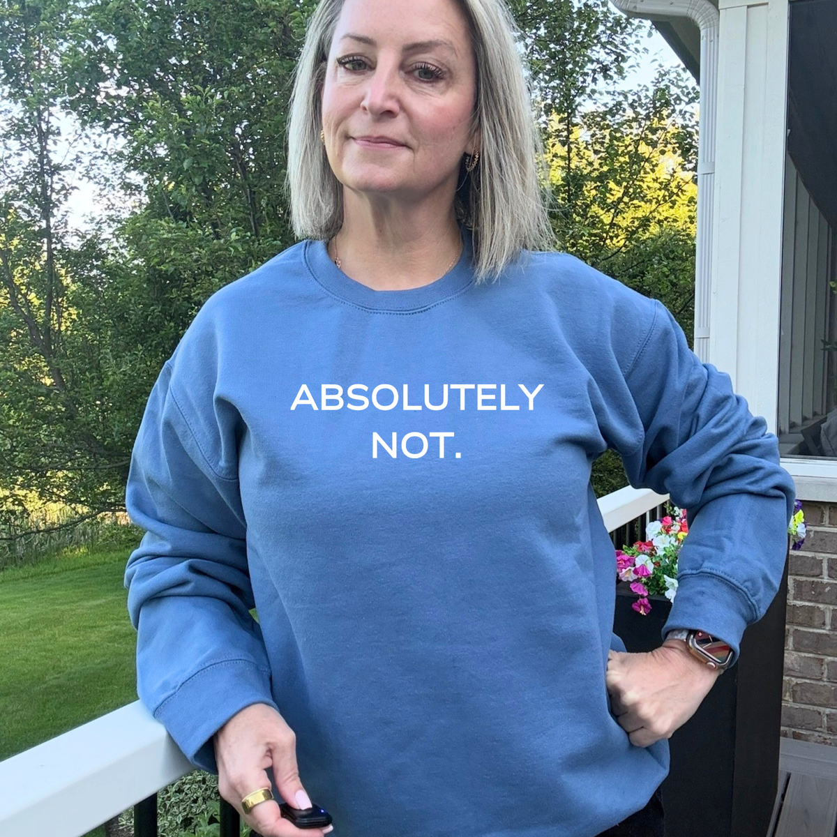 A woman wearing a dusty blue crewneck sweatshirt with the text 'ABSOLUTELY NOT.' printed in white across the chest, standing on a porch with trees in the background.