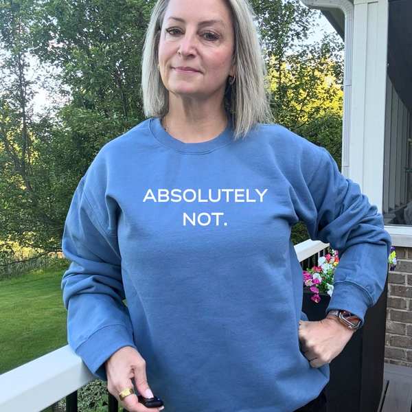A woman wearing a dusty blue crewneck sweatshirt with the text 'ABSOLUTELY NOT.' printed in white across the chest, standing on a porch with trees in the background.