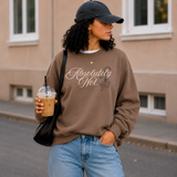 A woman wearing a brown sweatshirt with the text 'Absolutely Not.' in white script, paired with a black baseball cap, blue jeans, and carrying a black shoulder bag while holding an iced coffee on a city street.
