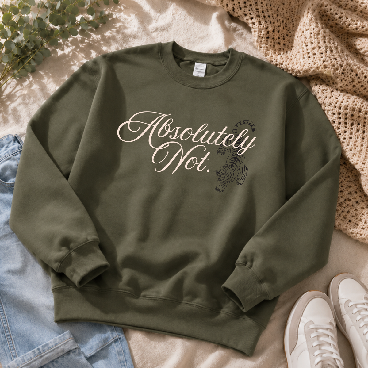 An olive green crewneck sweatshirt with 'Absolutely Not.' in white script and a tiger graphic, styled in a flat lay with jeans, a knit blanket, and sneakers.
