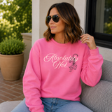 A woman wearing a bright pink crewneck sweatshirt with 'Absolutely Not.' printed in white script and a small tiger graphic, sitting on a gray couch outdoors.