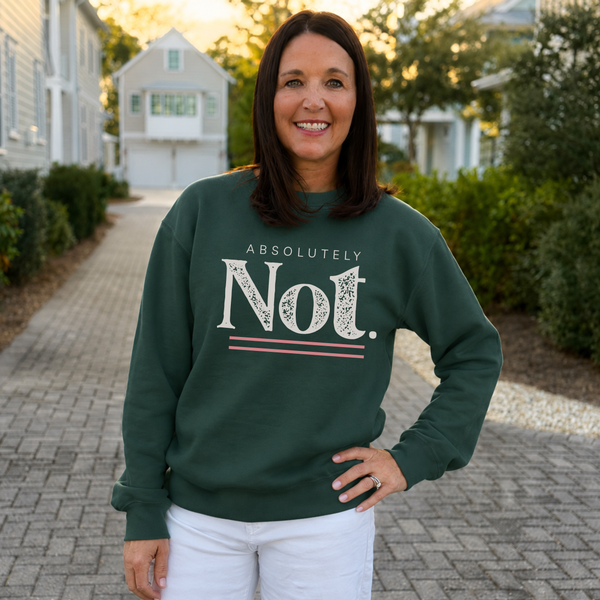 A woman stands on a paved walkway outdoors wearing a dark green crewneck sweatshirt with a white graphic print that reads 'ABSOLUTELY Not.' and white pants.