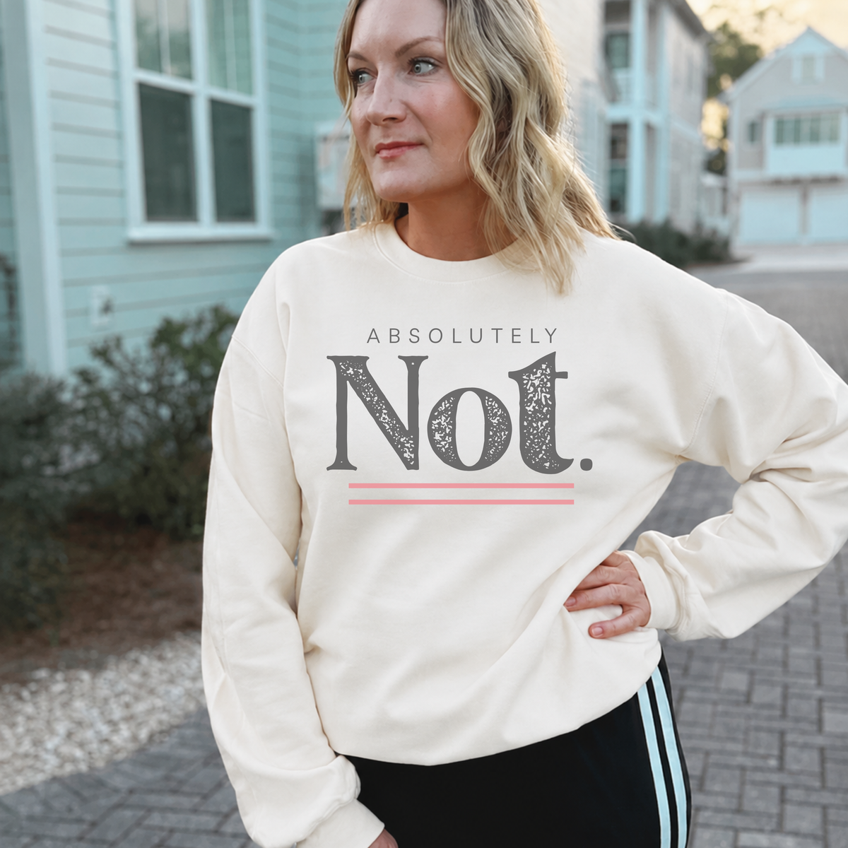 A woman wearing a cream-colored crewneck sweatshirt with a graphic print that reads 'ABSOLUTELY Not.' in dark gray with a pink underline accent, standing outdoors on a paved walkway with houses in the background.
