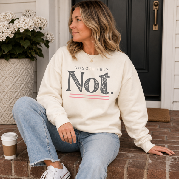 A woman sits on brick steps in front of a house door wearing a cream sweatshirt with a bold graphic that reads 'ABSOLUTELY Not.' in dark gray, with thin pink accent lines.