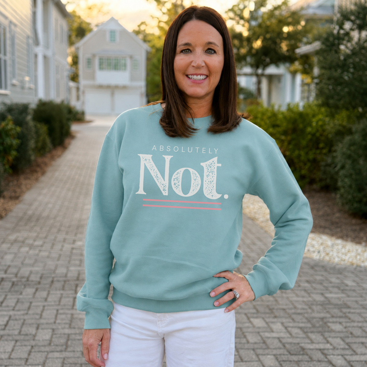 A woman standing on a paved walkway outdoors, wearing a light teal crewneck sweatshirt with a white graphic that reads 'ABSOLUTELY Not.' and white pants.