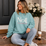 A woman sits on brick steps wearing a mint green sweatshirt with the text 'ABSOLUTELY Not.' in white and a thin coral line.