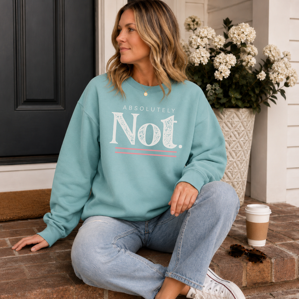 A woman sits on brick steps wearing a mint green sweatshirt with the text 'ABSOLUTELY Not.' in white and a thin coral line.