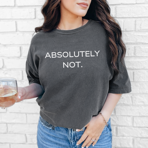 Woman wearing a dark gray t-shirt with 'ABSOLUTELY NOT.' text, holding a glass of white wine against a light brick wall.