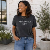 A woman wearing a dark charcoal gray t-shirt with the text 'ABSOLUTELY NOT.' printed in white across the chest, paired with blue jeans, standing in front of a white brick wall with potted plants.