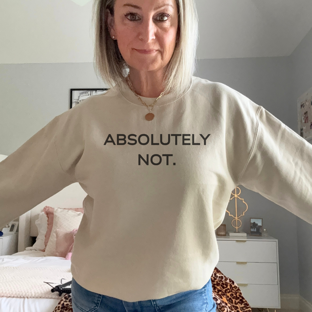 A woman wearing a cream-colored crewneck sweatshirt with the text 'ABSOLUTELY NOT.' printed in black across the chest, paired with blue jeans in a bedroom setting.