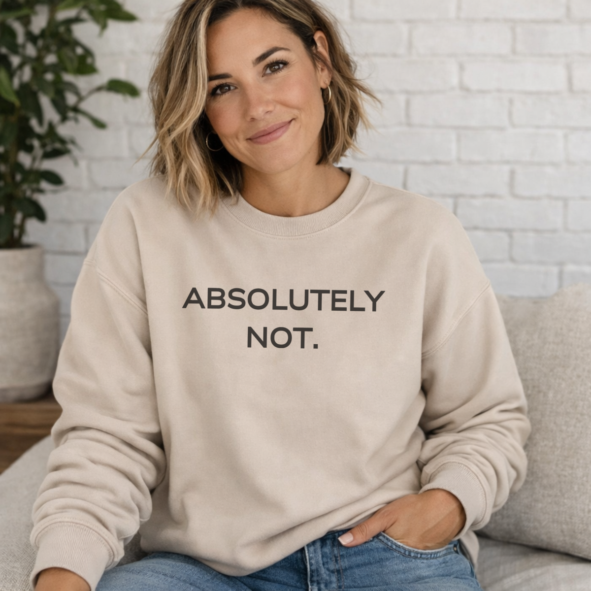 A woman wearing a beige crewneck sweatshirt with the text 'ABSOLUTELY NOT.' printed in black across the chest, sitting on a couch in front of a white brick wall.