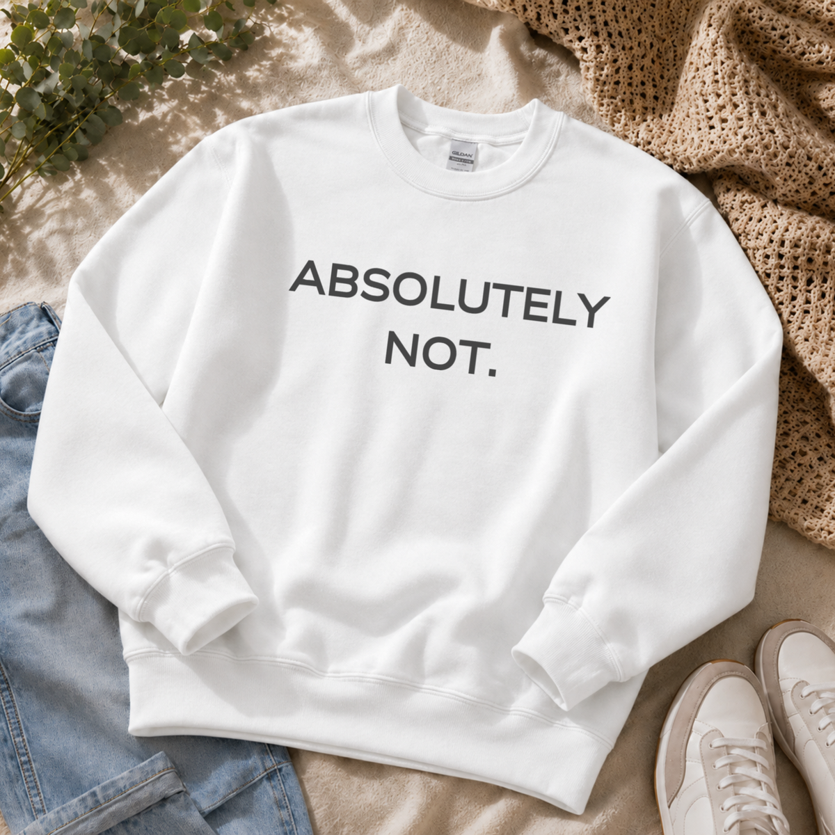 A white crewneck sweatshirt with the text 'ABSOLUTELY NOT.' printed in black across the chest, styled in a flat lay with blue jeans, a beige knit blanket, white sneakers, and green leaves.