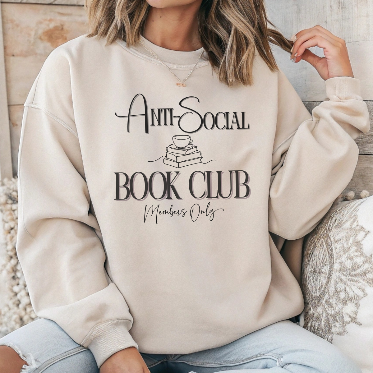 A woman wearing a cream-colored sweatshirt with a black graphic that reads 'Anti-Social Book Club Members Only' with a small book and cup illustration, paired with light blue jeans in a cozy indoor setting.