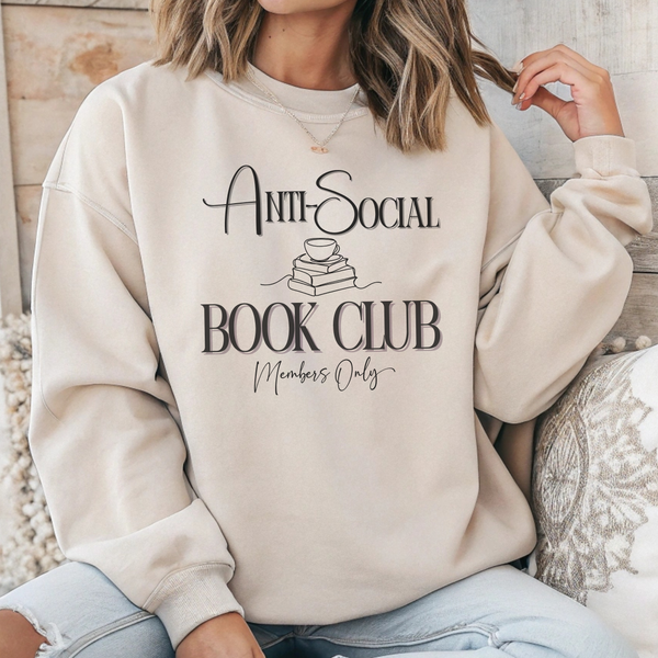 A woman wearing a cream-colored sweatshirt with a black graphic that reads 'Anti-Social Book Club Members Only' with a small book and cup illustration, paired with light blue jeans in a cozy indoor setting.