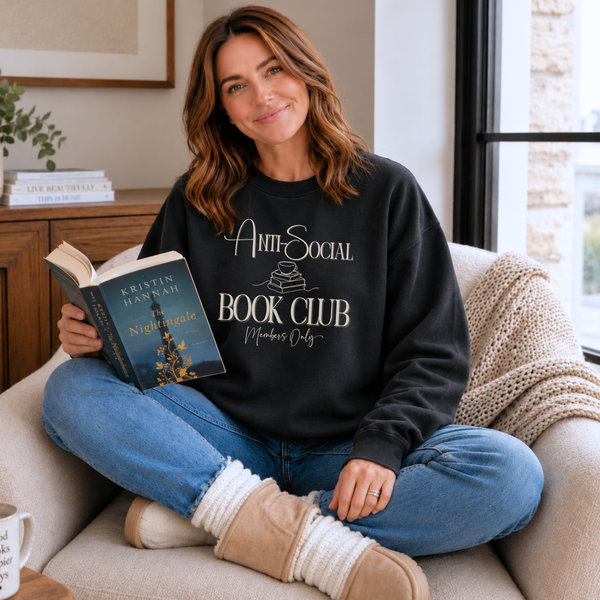 A woman sits cross-legged on a beige couch in a cozy living room, wearing a black crewneck sweatshirt with a white graphic that reads 'AntiSocial Book Club Members Only' while holding an open book.