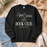 A black crewneck sweatshirt with a cream-colored graphic reading 'ANTI-SOCIAL BOOK CLUB Members Only' laid flat on a beige fabric background, styled with blue jeans, white sneakers, a knit blanket, and greenery.