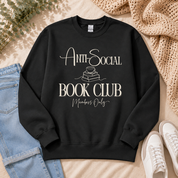 A black crewneck sweatshirt with a cream-colored graphic reading 'ANTI-SOCIAL BOOK CLUB Members Only' laid flat on a beige fabric background, styled with blue jeans, white sneakers, a knit blanket, and greenery.