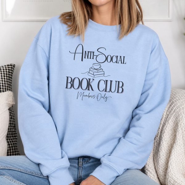 A person wearing a light blue crewneck sweatshirt with a black graphic that reads 'Anti-Social Book Club Members Only' sitting on a couch.
