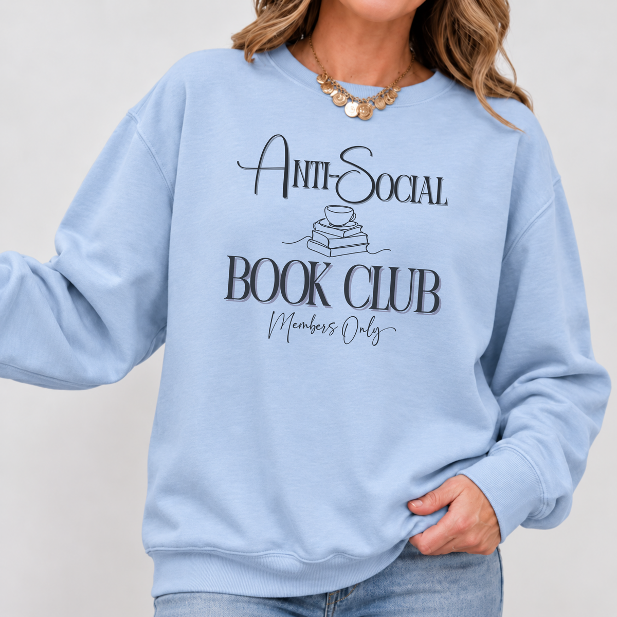 A woman wearing a light blue crewneck sweatshirt with a black graphic that reads 'Anti-Social Book Club Members Only' with a small book illustration, paired with blue jeans and a gold necklace.