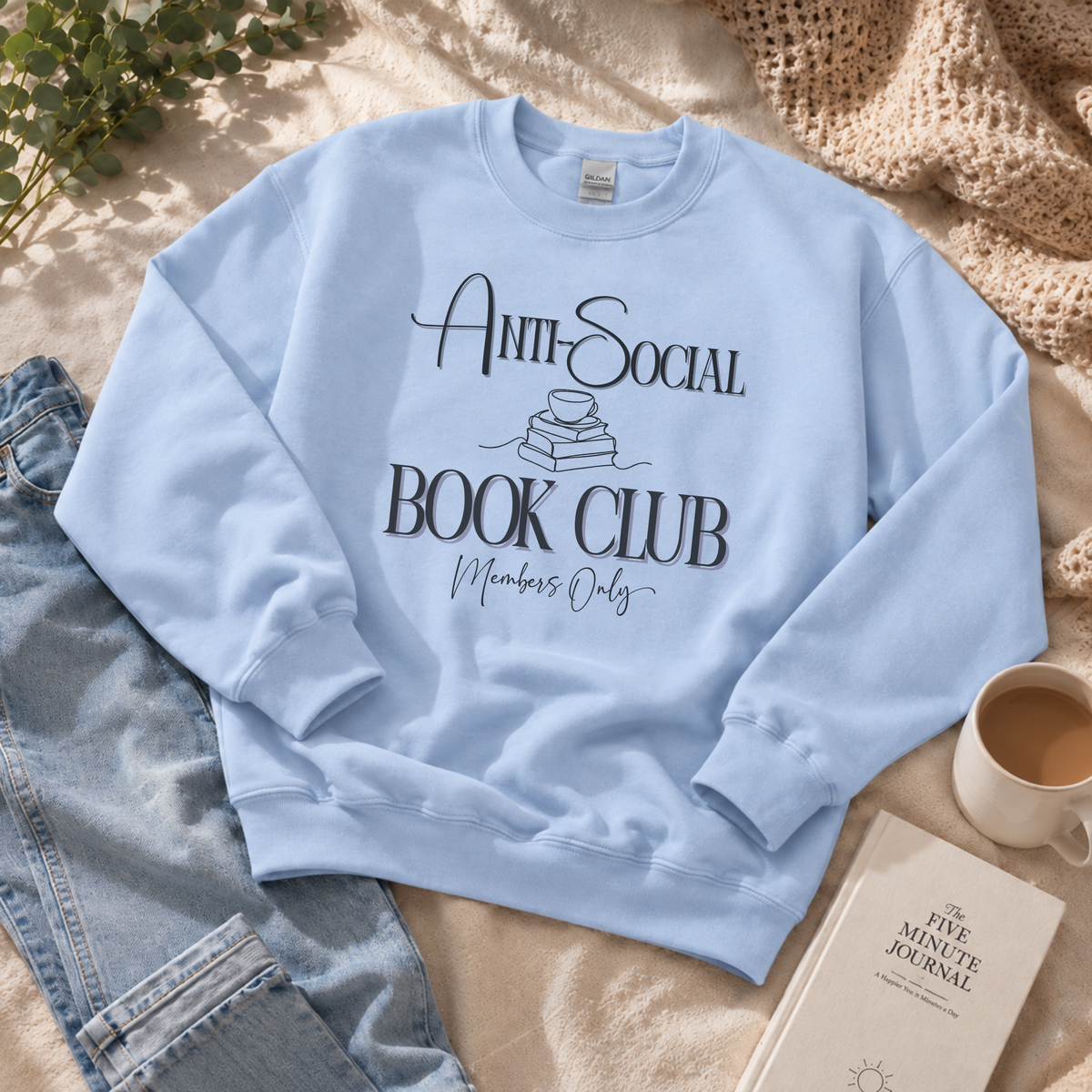 A light blue crewneck sweatshirt with a black graphic reading 'Anti-Social Book Club Members Only' laid flat on a beige blanket with jeans, a mug of coffee, and a journal nearby.