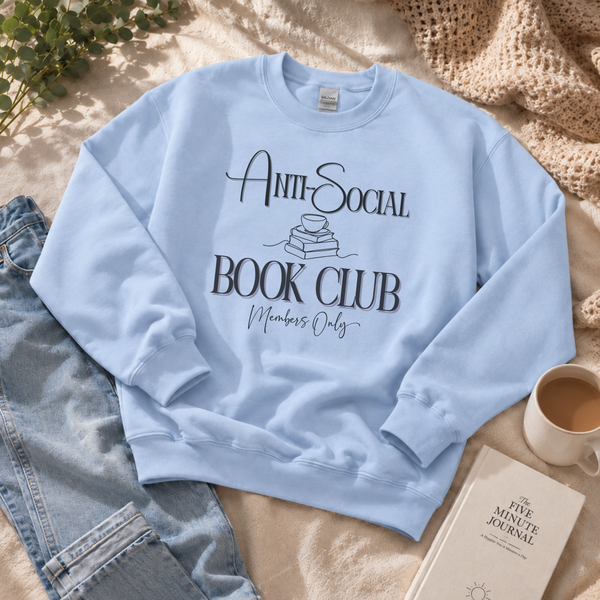 A light blue crewneck sweatshirt with a black graphic reading 'Anti-Social Book Club Members Only' laid flat on a beige blanket with jeans, a mug of coffee, and a journal nearby.