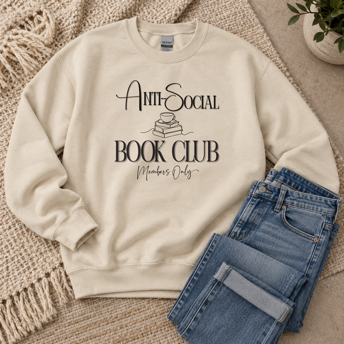 A cream-colored crewneck sweatshirt with a black graphic reading 'Anti-Social Book Club Members Only' laid flat on a woven rug next to folded blue jeans.