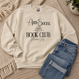 A cream-colored crewneck sweatshirt with a black graphic reading 'Anti-Social Book Club Members Only' laid flat on a woven rug next to folded blue jeans.