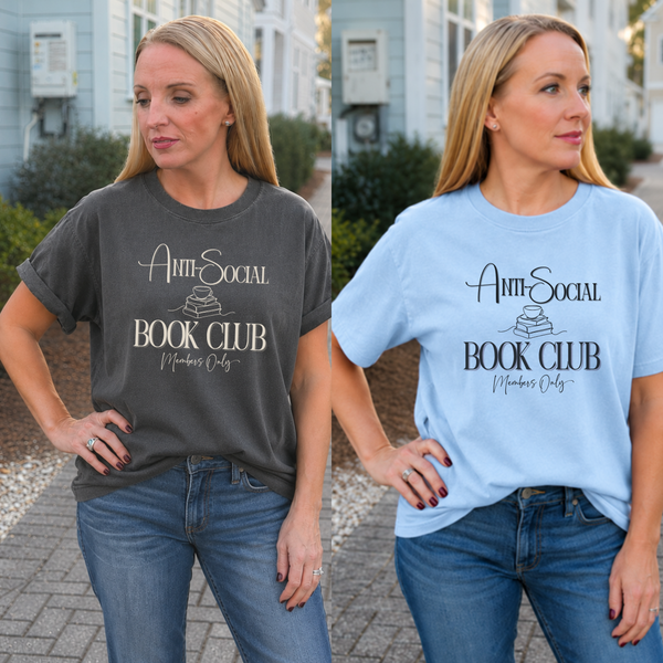 Two women standing outdoors side by side, each wearing a graphic T-shirt. The left shirt is dark charcoal gray with white text reading 'Anti-Social Book Club Members Only,' and the right shirt is light blue with the same text in black.