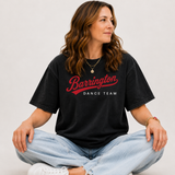 A woman sitting cross-legged wearing a faded black t-shirt with 'Barrington DANCE TEAM' printed in red and white across the chest, paired with light-wash jeans and white sneakers, against a plain light gray background.