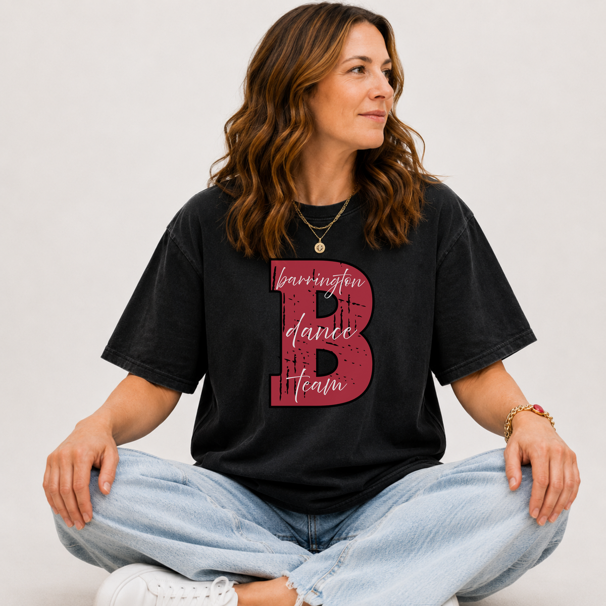 A woman sitting cross-legged wearing a faded black t-shirt with a large red graphic letter 'B' and the words 'barrington dance team' on the front, paired with light blue jeans and white sneakers against a light gray background.