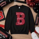 A black crewneck sweatshirt laid flat on a wood surface, featuring a large distressed red letter B with the words 'barrington dance team' in white script across it. Surrounding props include a black and red cheer pom, a water bottle, white sneakers, and sports bags with Barrington Broncs branding.