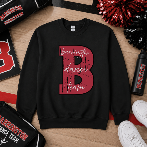 A black crewneck sweatshirt laid flat on a wood surface, featuring a large distressed red letter B with the words 'barrington dance team' in white script across it. Surrounding props include a black and red cheer pom, a water bottle, white sneakers, and sports bags with Barrington Broncs branding.
