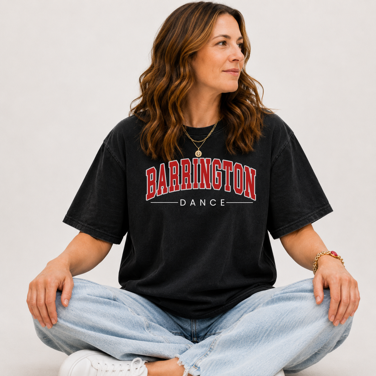 A woman sitting cross-legged wearing a faded black graphic t-shirt with 'BARRINGTON DANCE' printed in red and white, paired with light wash jeans and white sneakers, against a plain light gray background.