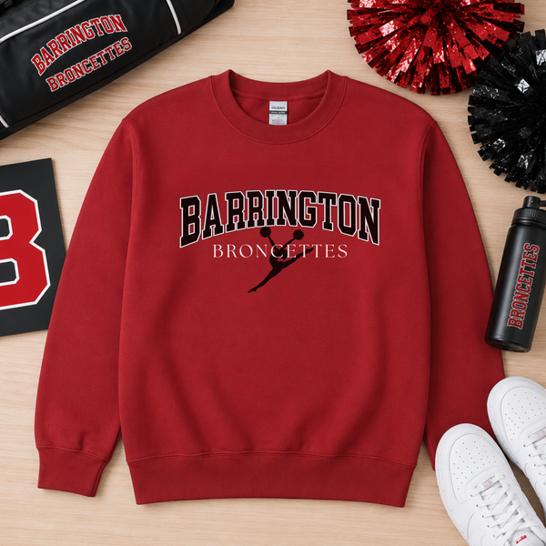 A red crewneck sweatshirt with 'BARRINGTON BRONCETTES' printed on the front, styled in a flat lay on a light wood background with sports-themed props including a black bag, cheer pom-poms, a water bottle, and white sneakers.