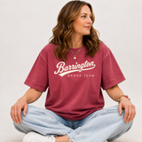 A woman sitting cross-legged wearing a faded burgundy t-shirt with 'Barrington Dance Team' printed in white across the chest, paired with light-wash jeans and white sneakers, against a plain light gray background.