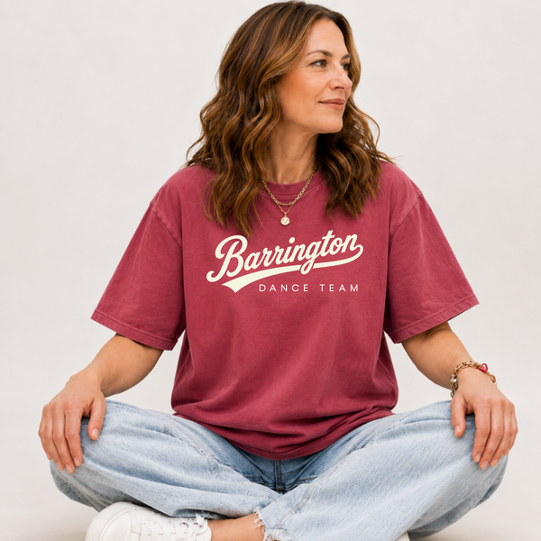 A woman sitting cross-legged wearing a faded burgundy t-shirt with 'Barrington Dance Team' printed in white across the chest, paired with light-wash jeans and white sneakers, against a plain light gray background.