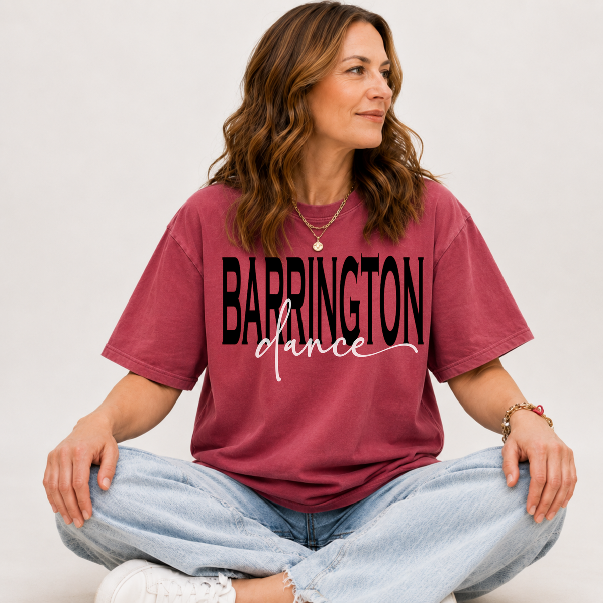 A woman sitting cross-legged wearing a faded burgundy t-shirt with 'BARRINGTON dance' printed in black across the front, paired with light-wash jeans and white sneakers, against a plain light gray background.