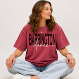 A woman sitting cross-legged wearing a faded burgundy t-shirt with 'BARRINGTON dance' printed in black across the front, paired with light-wash jeans and white sneakers, against a plain light gray background.
