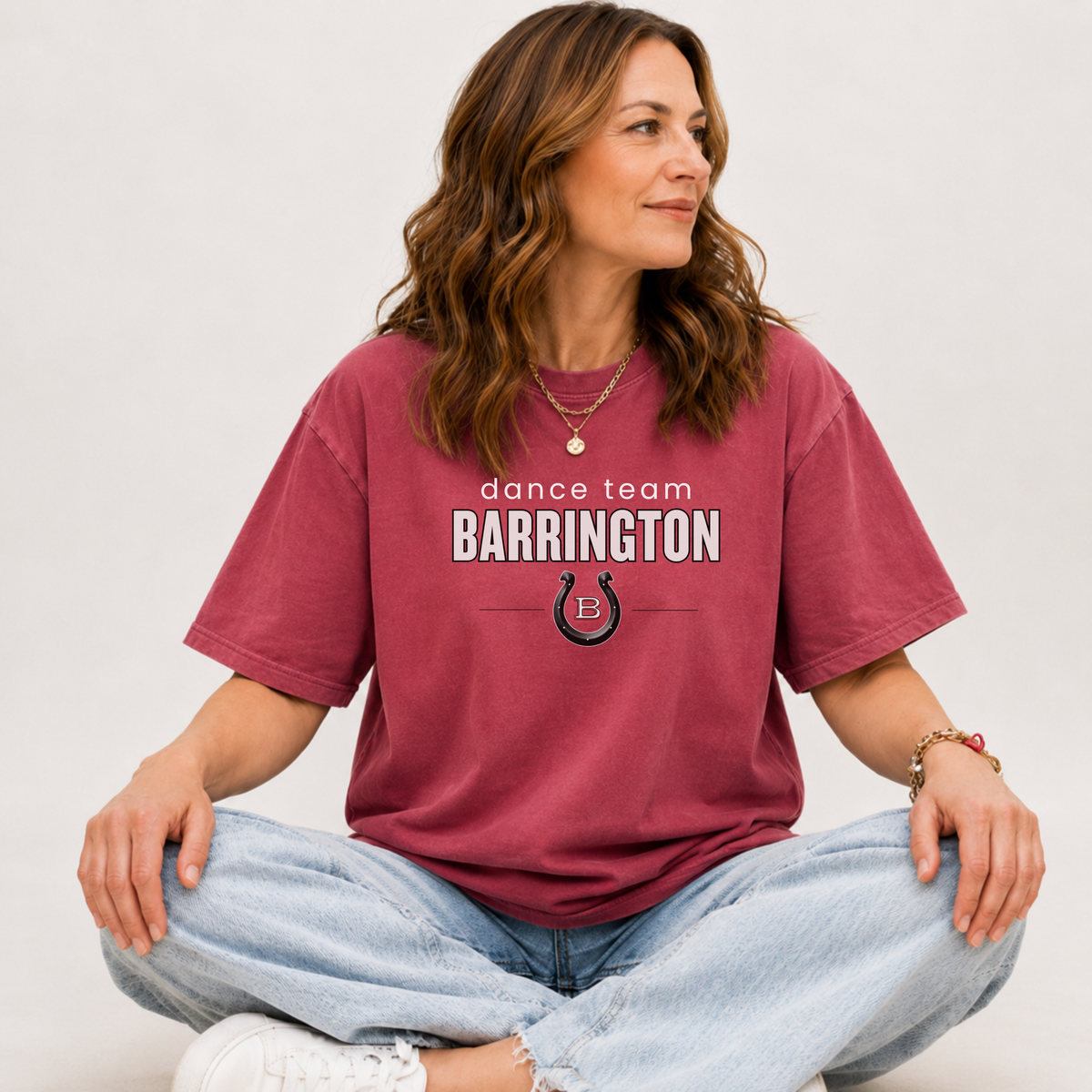 A woman sitting cross-legged wearing a maroon short-sleeve graphic T-shirt that reads 'dance team BARRINGTON' with a horseshoe logo, paired with light blue jeans and white sneakers, against a plain light gray background.